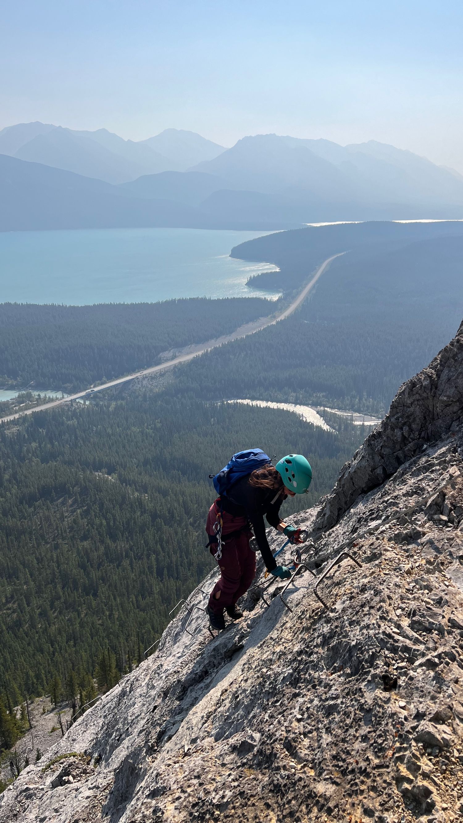 A climber descends a steep rocky slope overlooking a winding mountain road and turquoise lake surrounded by forested peaks. Kristen is cool