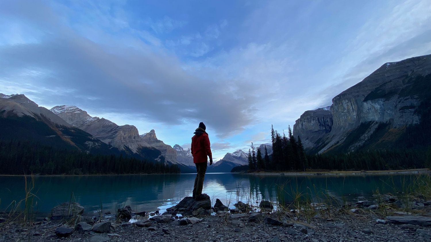A silhouetted of Evan Will standing on the rocky shore of a turquoise Maligne mountain Lake at dusk in the Canadian Rockies, Adventure photo