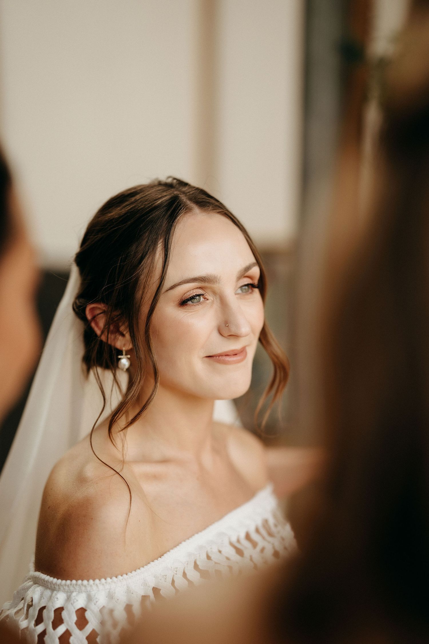 A beautiful bride in an off-shoulder lace wedding dress smiles warmly while getting ready for her special day.