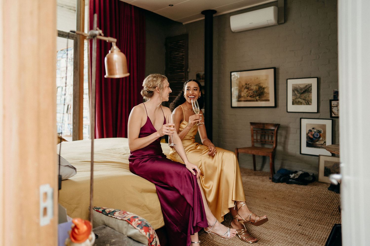Two people in elegant dresses sit together in a cozy room decorated with framed artwork and burgundy curtains.