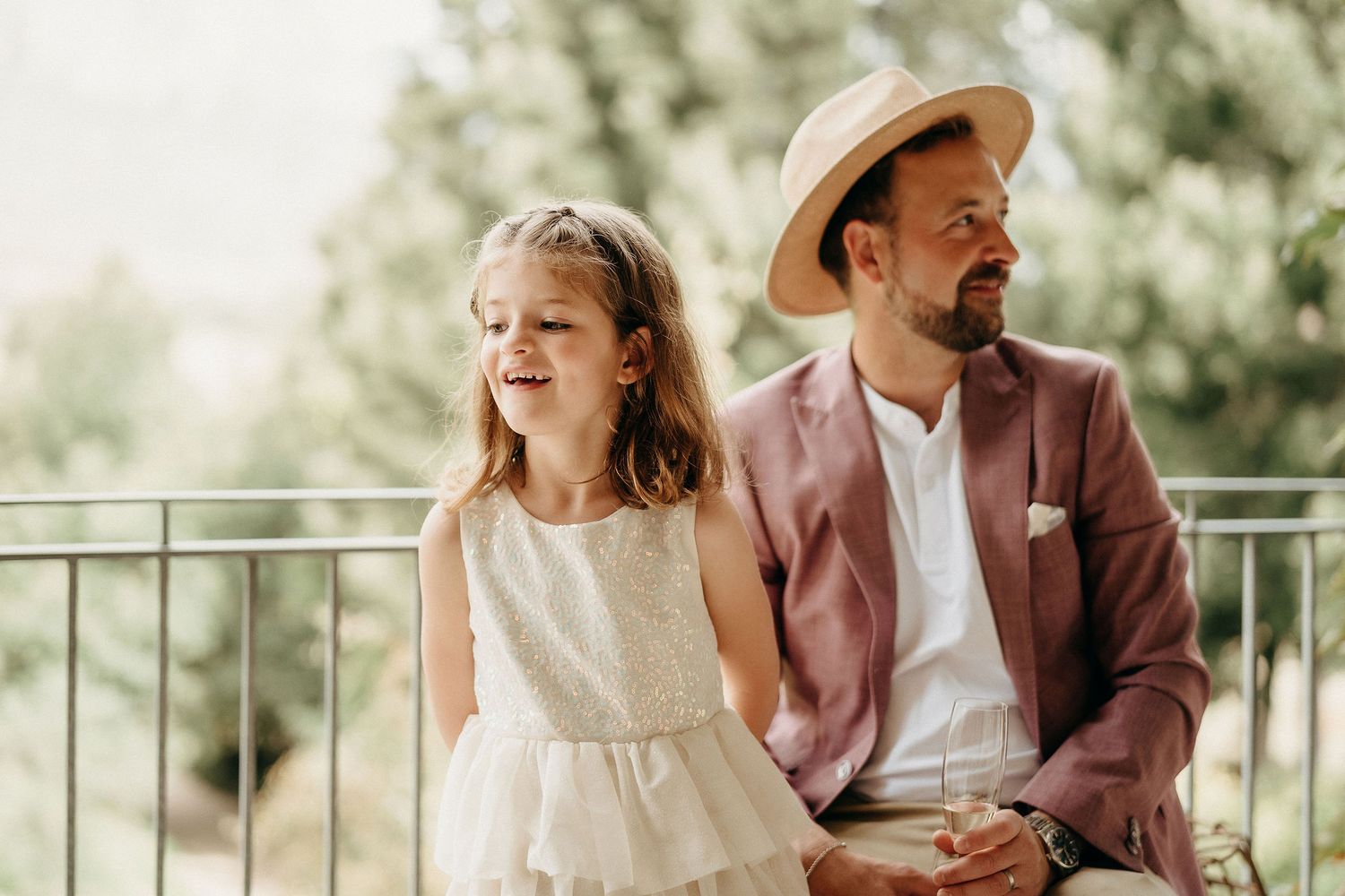 A parent and child sit together on a balcony, dressed in summer attire with a pink blazer and white dress.