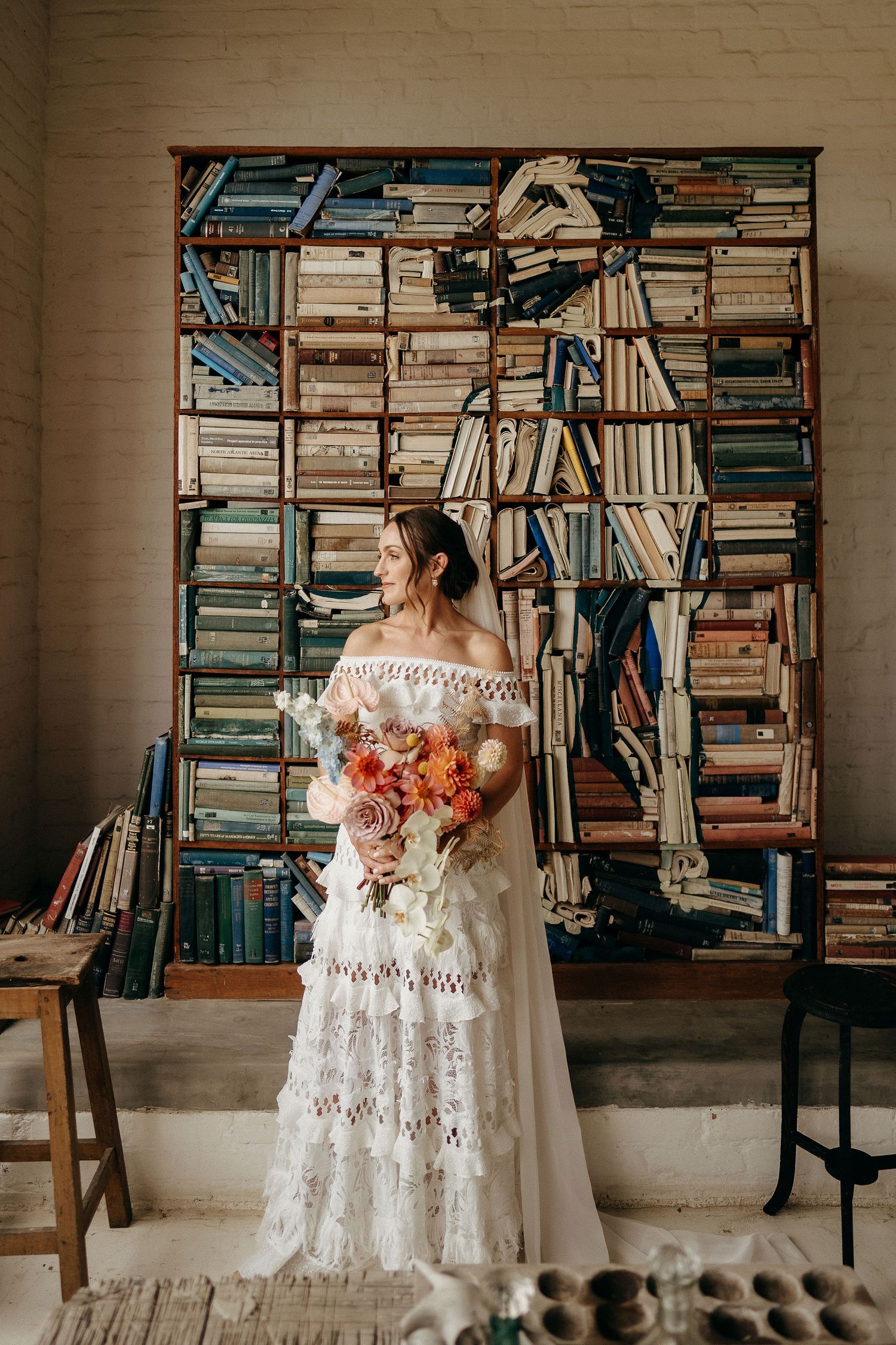 A woman in a white lace wedding dress holds a vibrant coral bouquet while standing against a wall of stacked books.