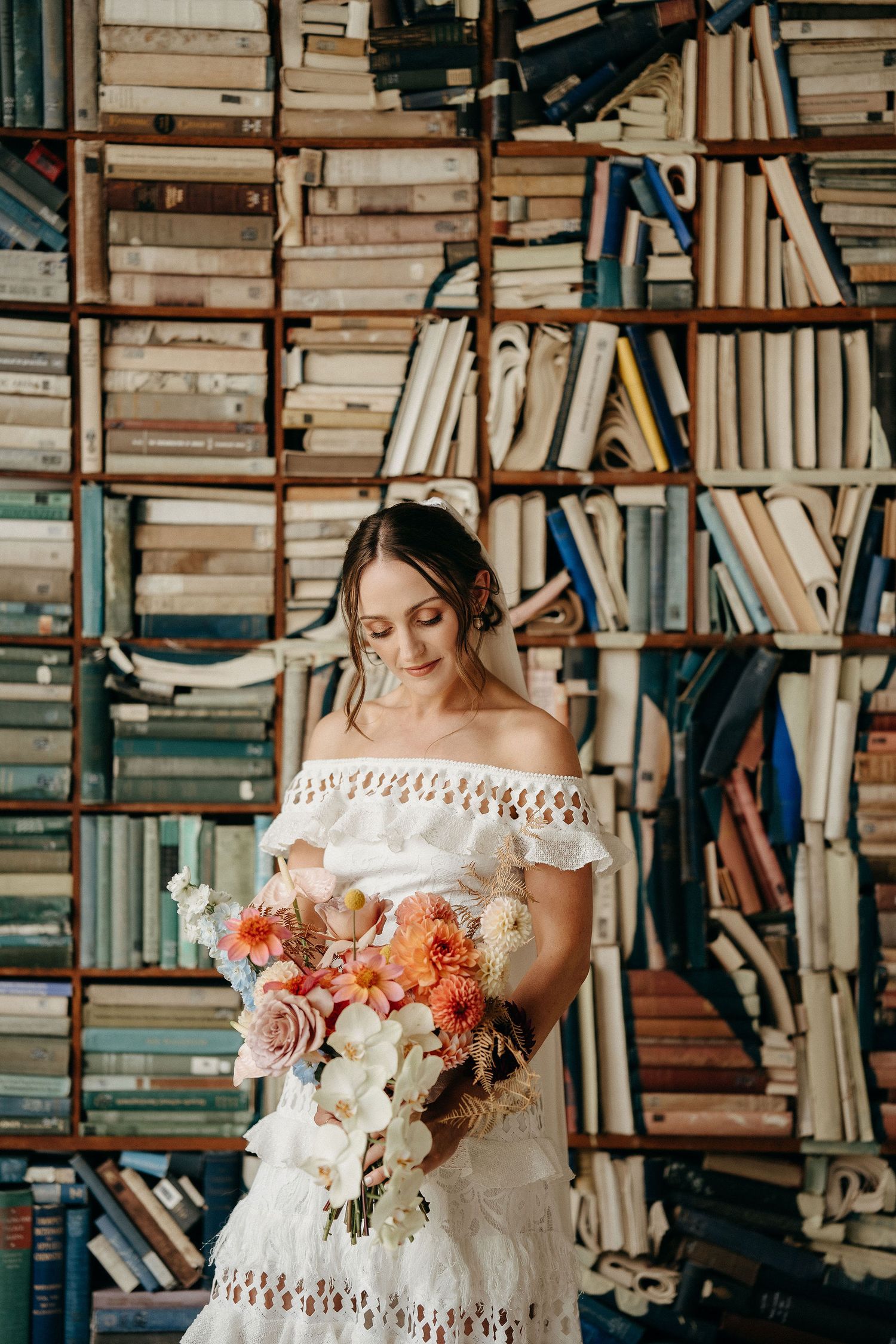 Romantic setting in library with off-shoulder white lace dress and colorful floral bouquet against vintage bookshelf backdrop.