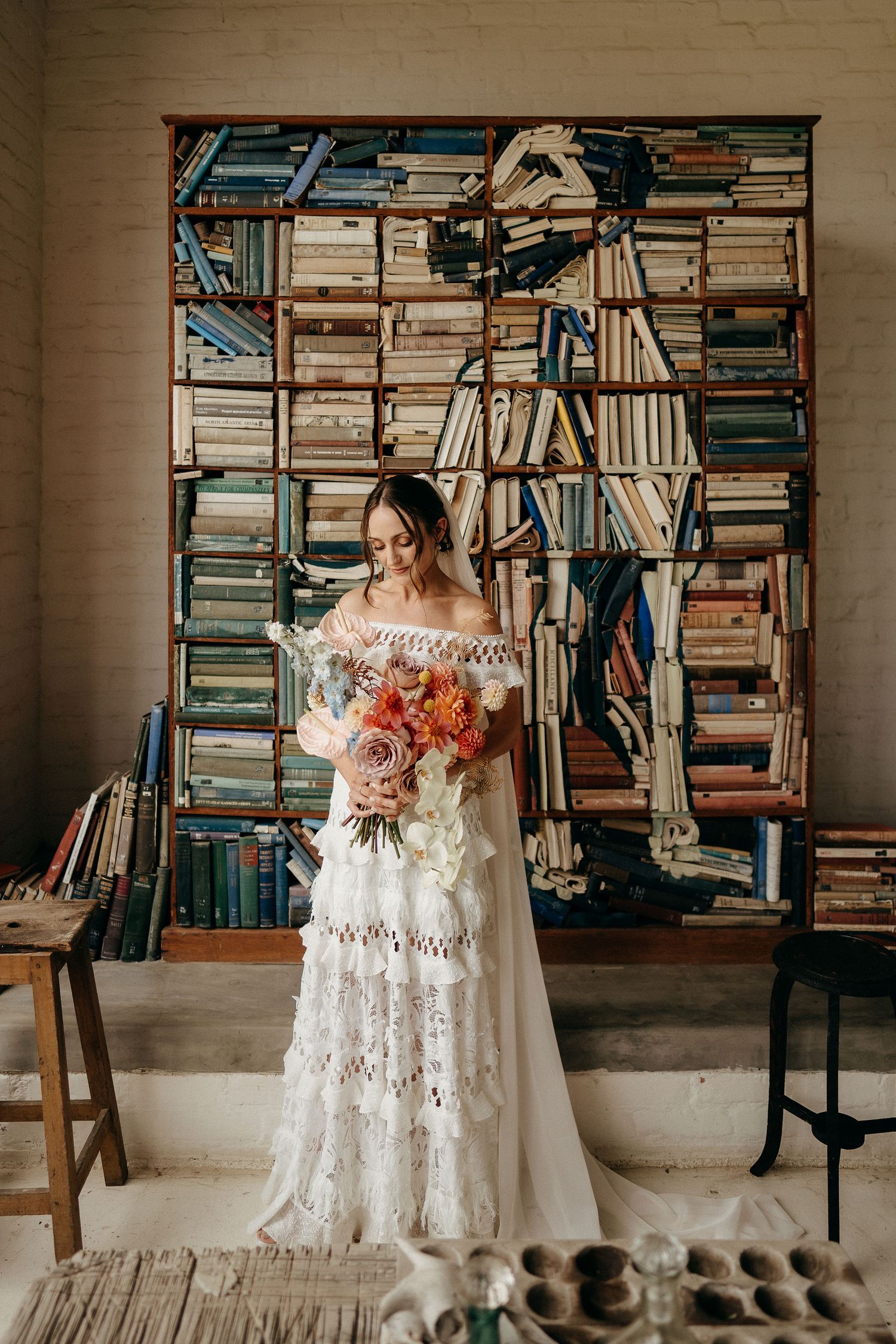A bride in a white lace gown holds a coral bouquet while standing in front of floor-to-ceiling bookshelves.