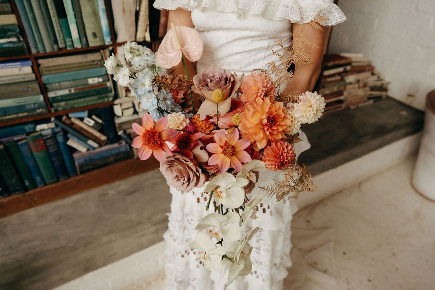 A romantic peach and coral wedding bouquet with mixed flowers held in front of a white lace gown.