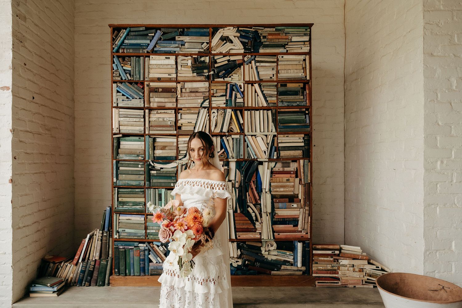 Person in white lace wedding dress holds vibrant coral bouquet while standing against wall of stacked vintage books.