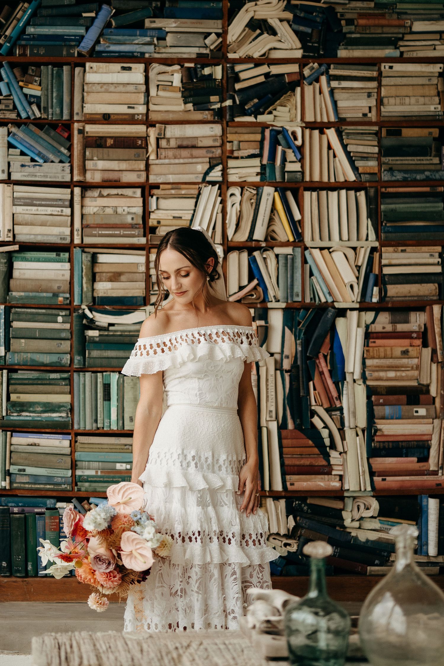 A bride in an off-shoulder lace dress stands before towering bookshelves holding a peach and white bouquet.