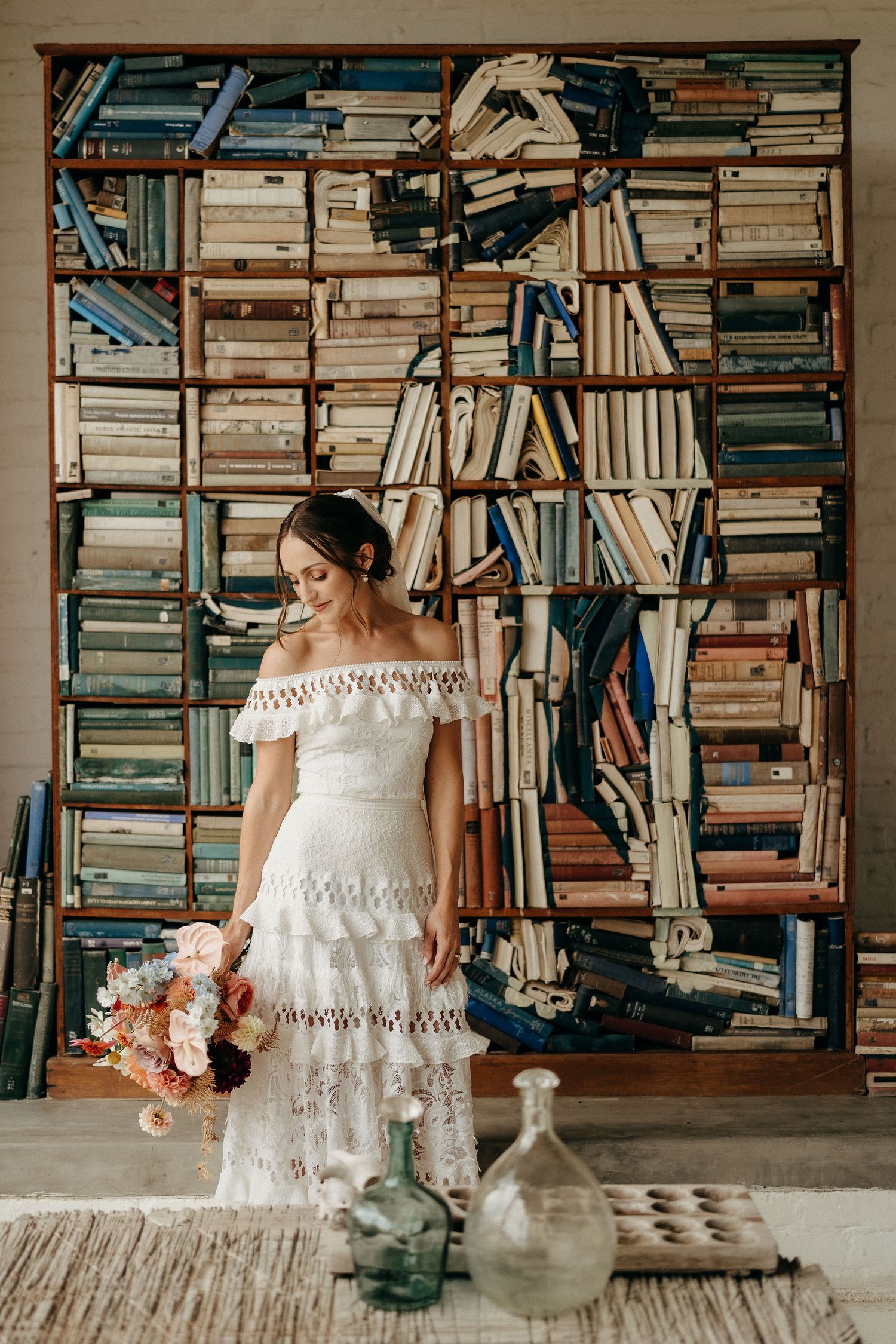Bride in layered off-shoulder dress poses against wall of vintage books with glass bottles on floor.