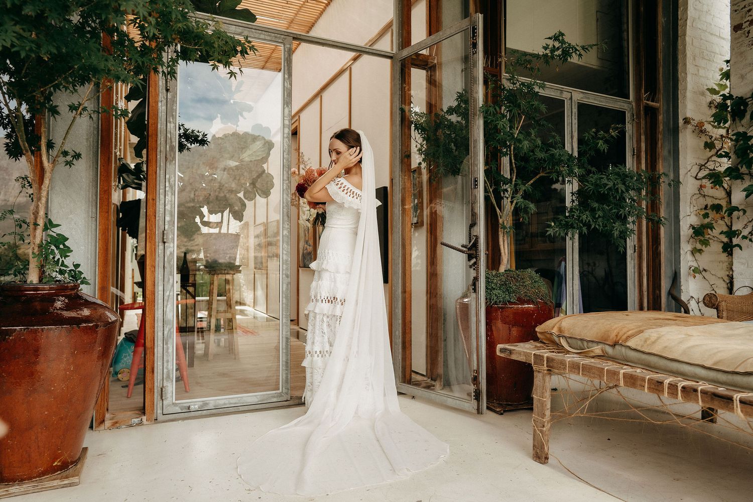Elegant bridal portrait in white dress standing by glass doors in sunlit greenhouse setting.