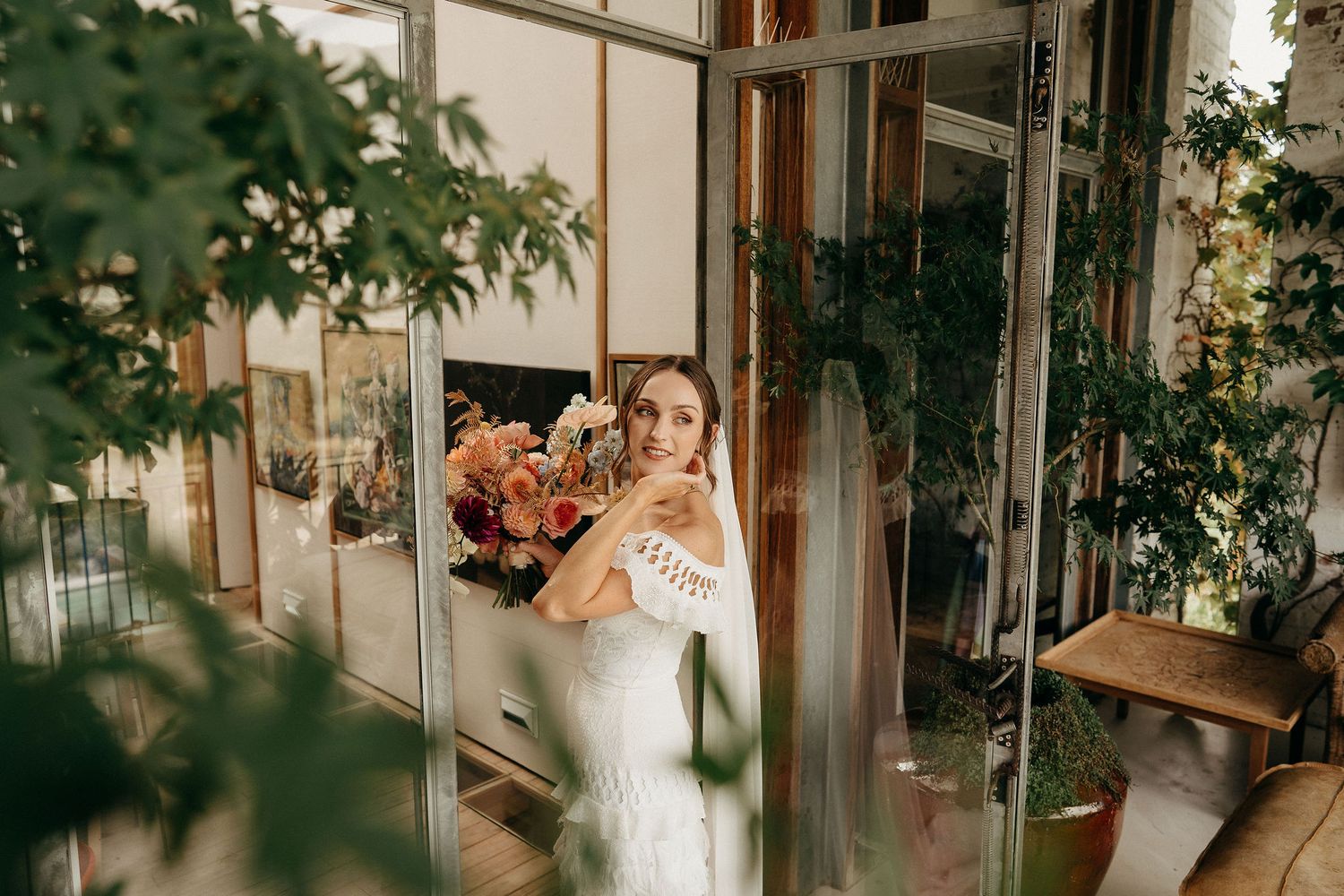 Sunlit bride holding coral bouquet poses in front of glass windows with green foliage.