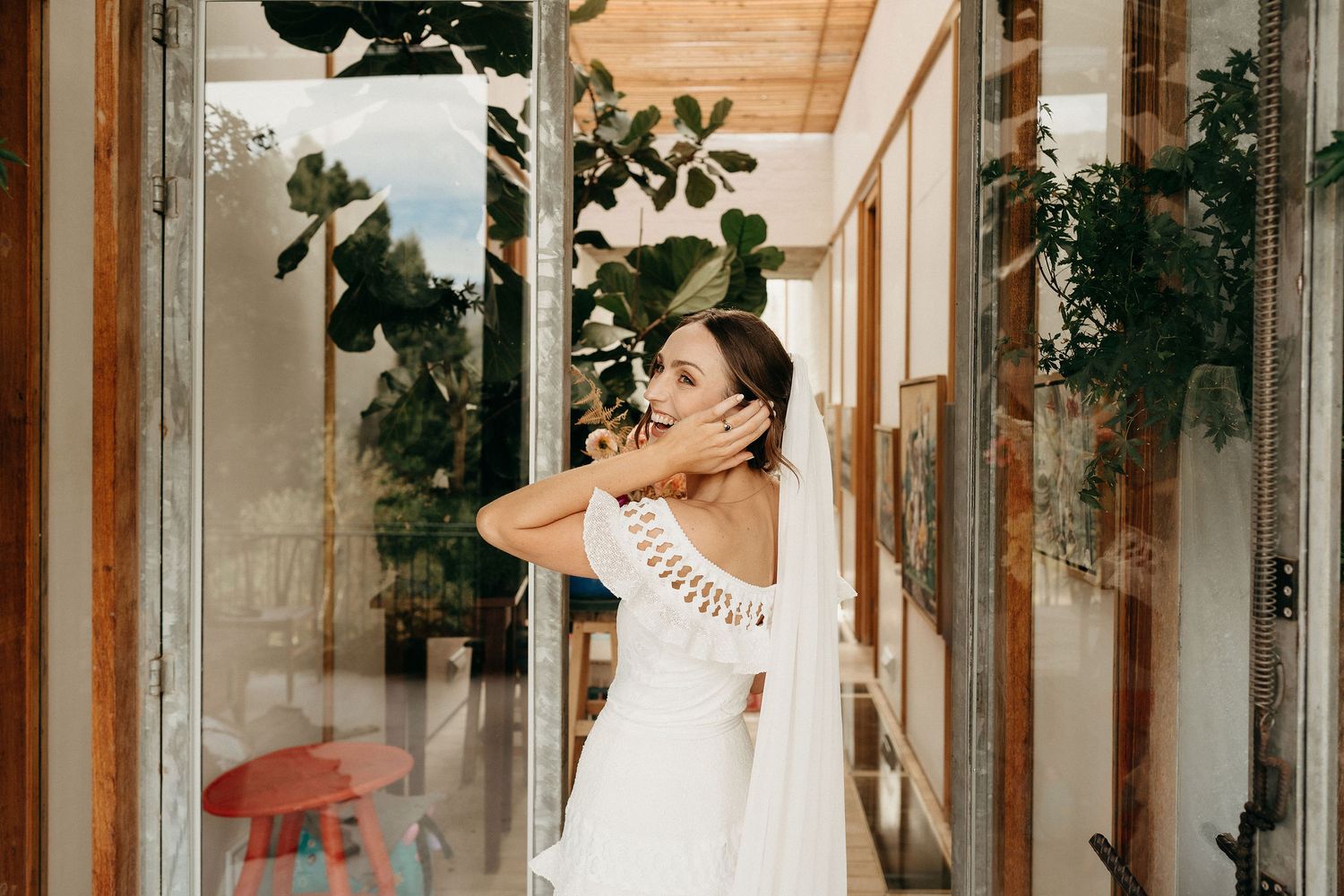 A figure in a white summer dress poses gracefully against wooden columns and greenery in a sunlit space.