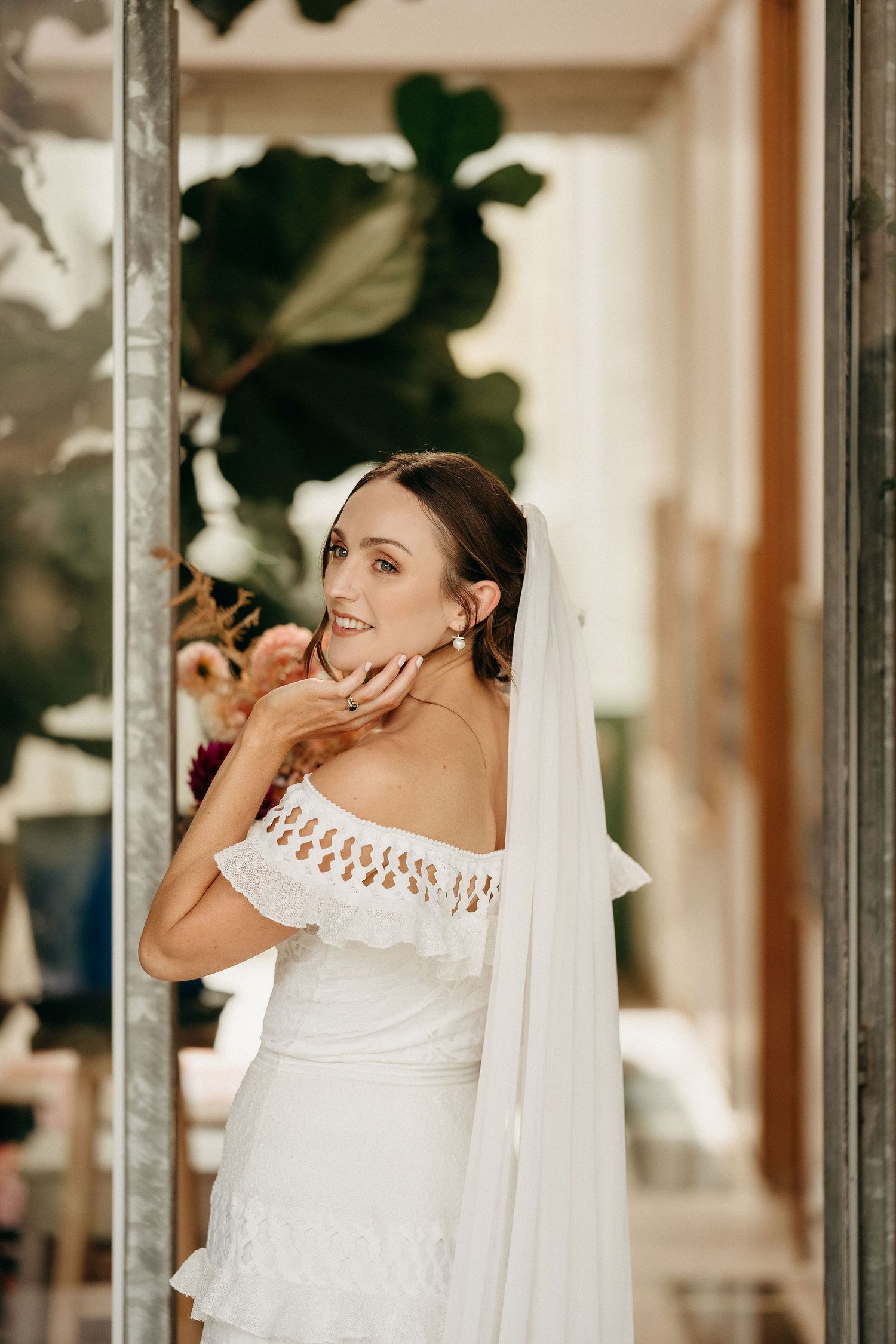 A bride in an elegant white off-shoulder wedding dress with lace details and flowing veil poses against a leafy backdrop.
