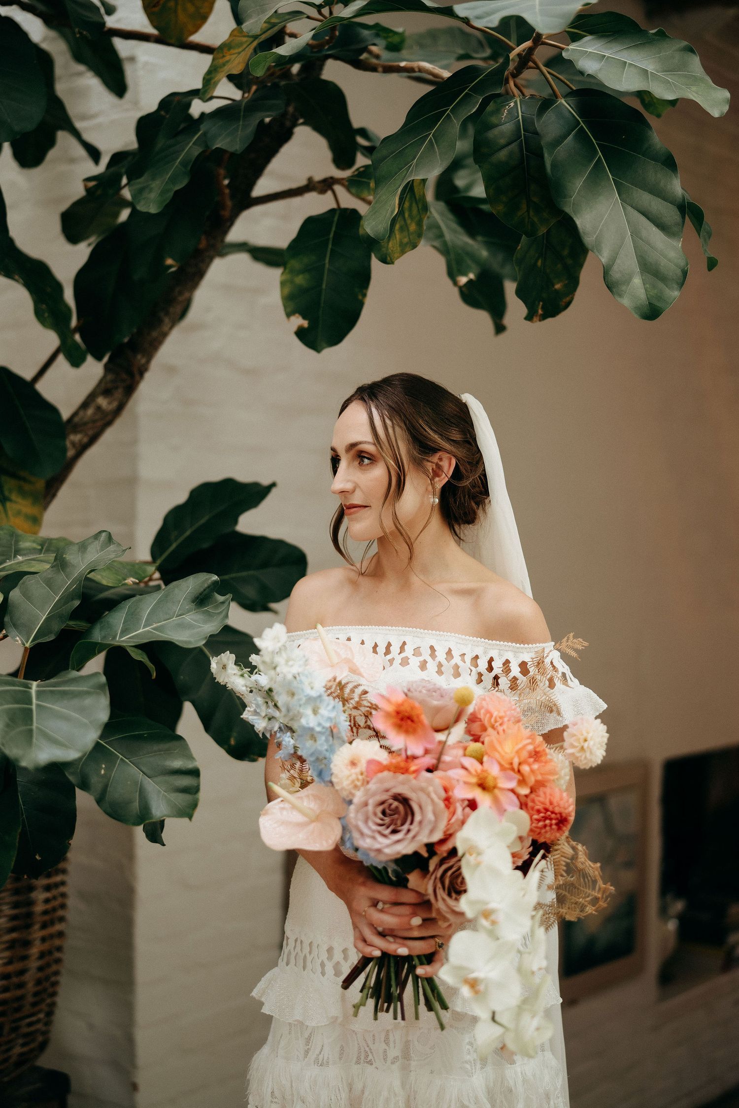 Elegant off-shoulder lace wedding dress with coral and peach bouquet under lush fiddle leaf fig tree branches.