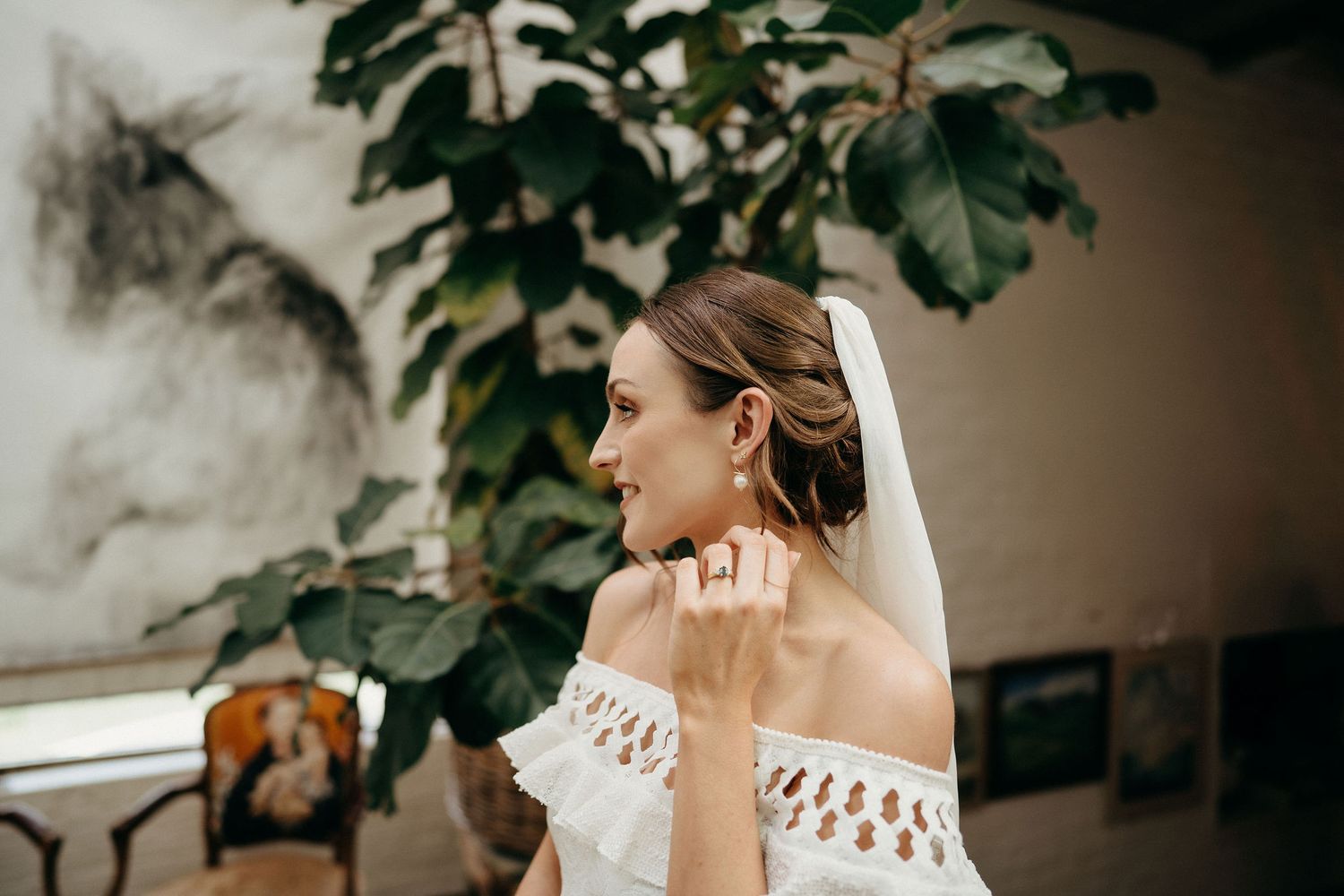 A bride in an elegant off-shoulder lace dress stands in front of green foliage in a dramatic side profile.