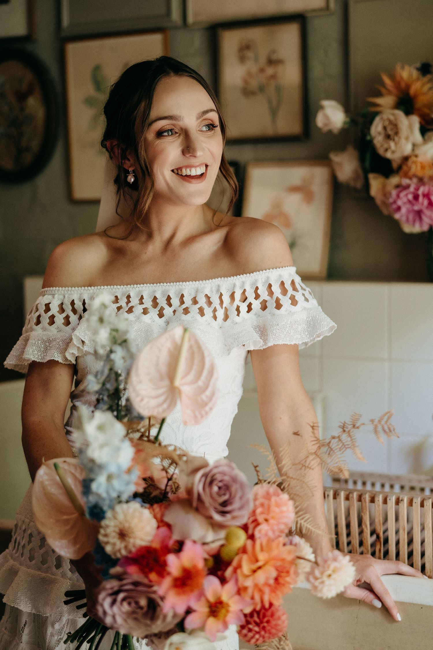 A bride in an off-shoulder lace gown holds a vibrant bouquet of pink, peach and coral flowers against a rustic backdrop.