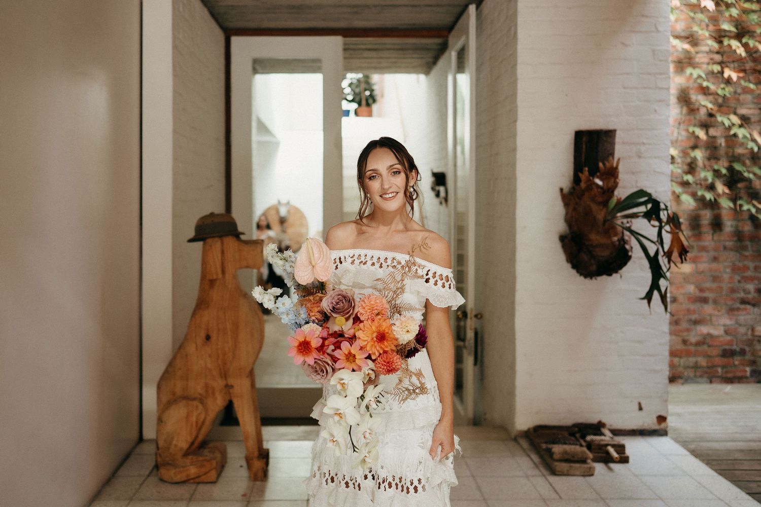A bride in a lace off-shoulder gown holds a vibrant coral and peach bouquet while standing in a rustic hallway.