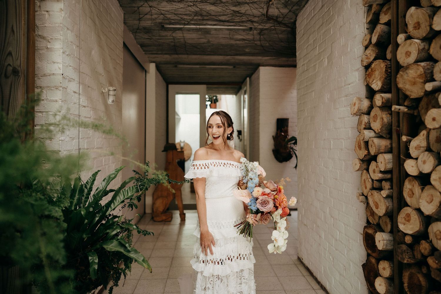 A person in a white off-shoulder wedding dress holds a bouquet while standing in a rustic hallway with stacked firewood.