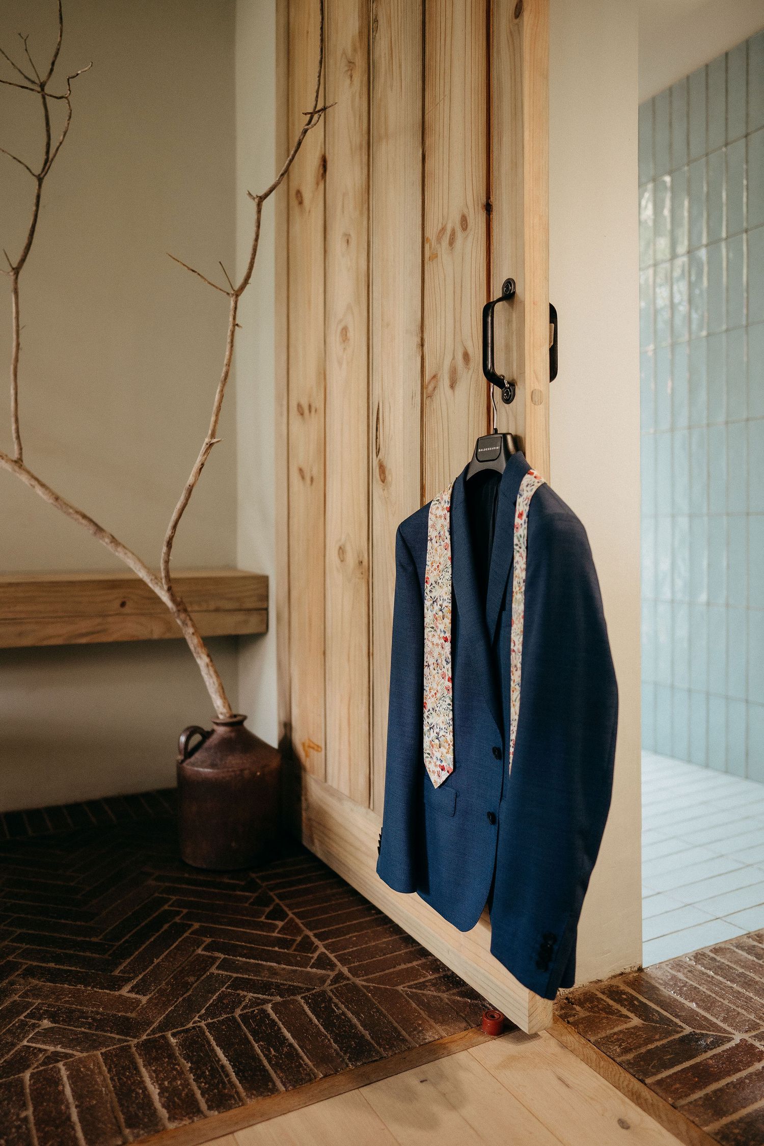 A navy blue suit jacket with floral tie hangs on a wooden door near a decorative branch in a rustic interior.