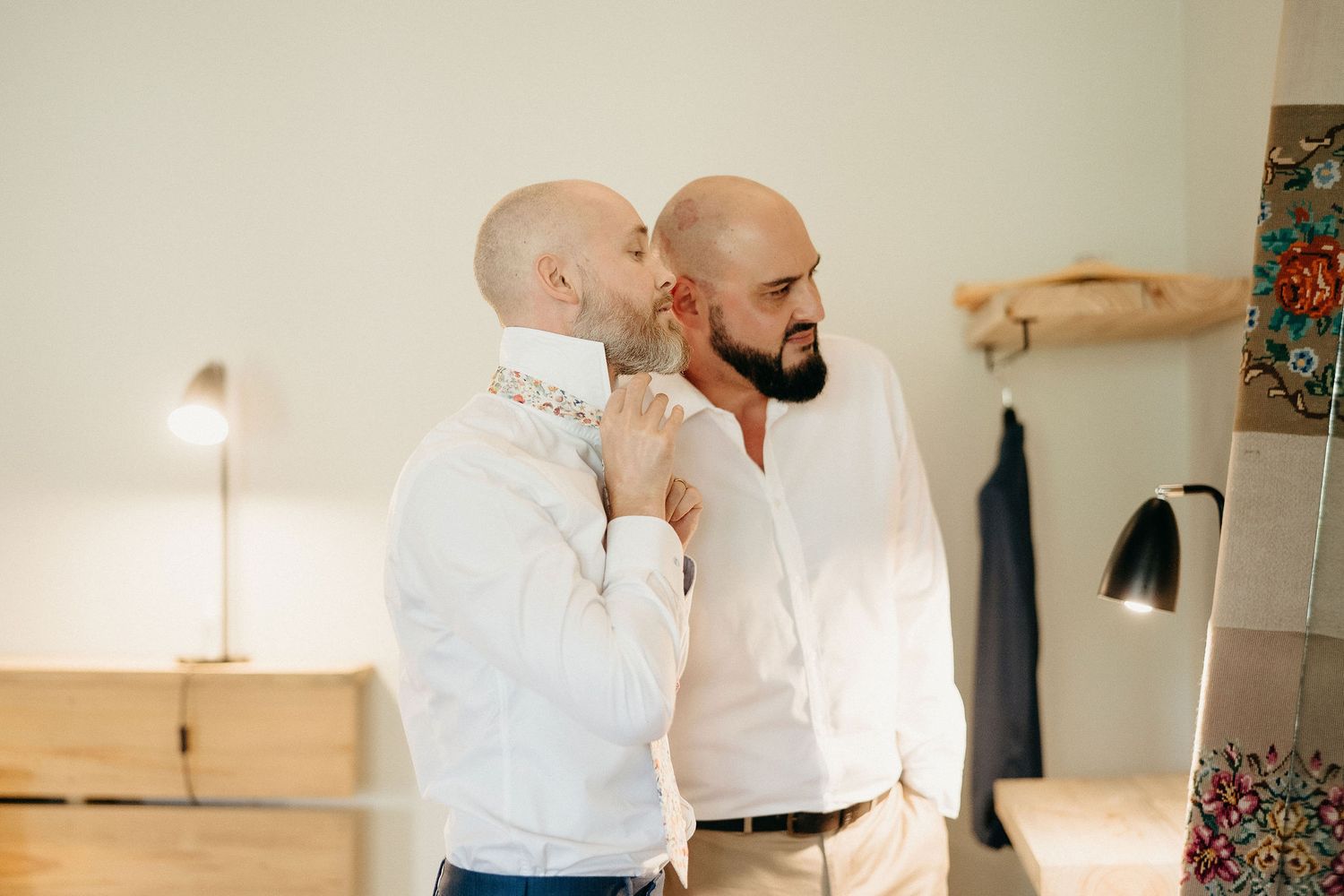 Two men in white dress shirts sharing a tender moment while getting dressed in a well-lit room.