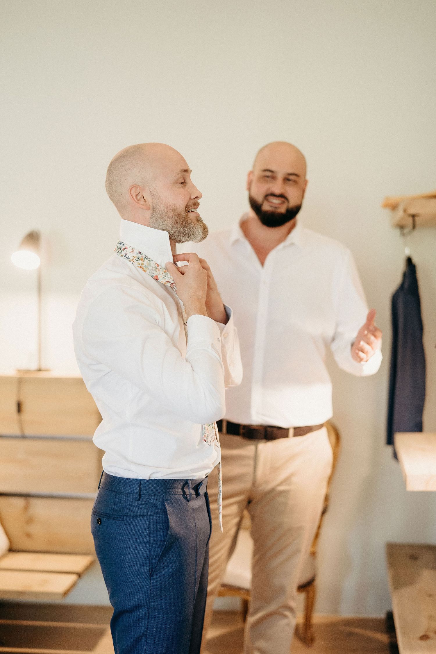 Two men in white shirts stand in a room, one fixing his bowtie while the other gestures in conversation.