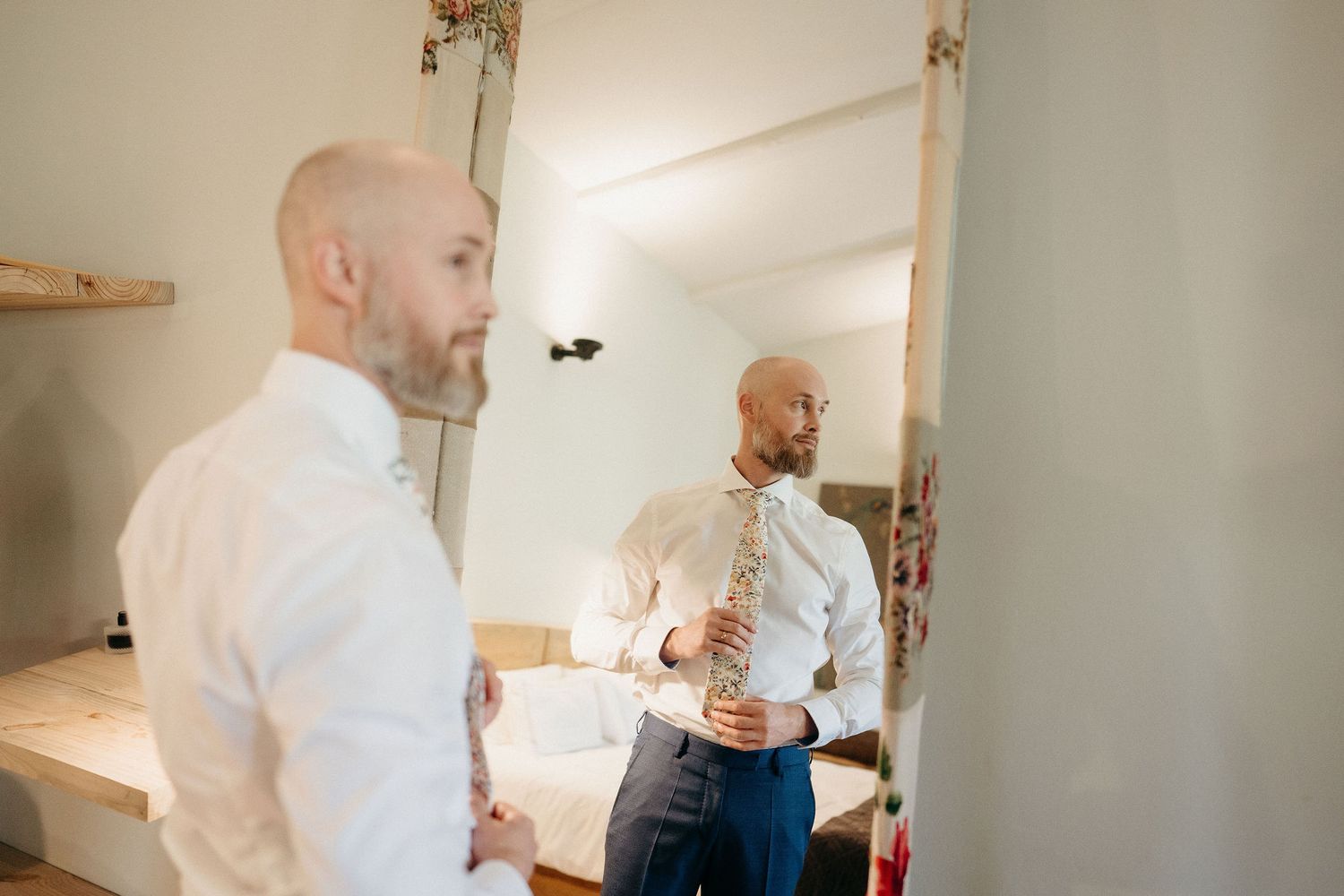 Person in white dress shirt adjusts tie while looking in mirror during preparation.