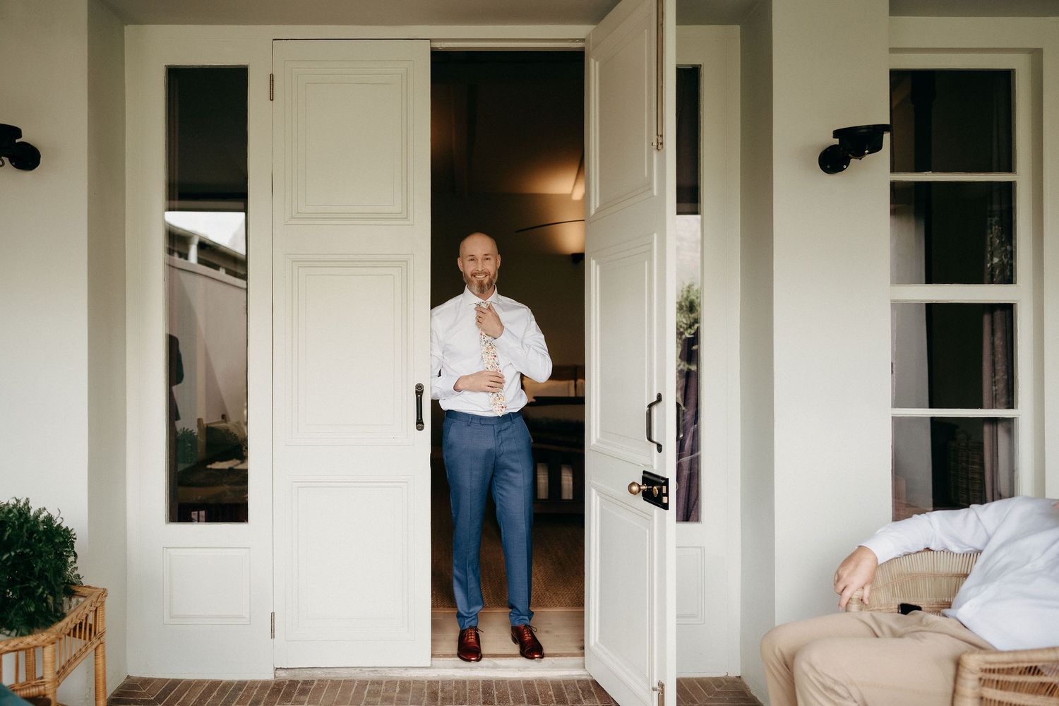 A casual scene shows someone in denim and a white shirt standing in a doorway of a white porch with outdoor furniture.