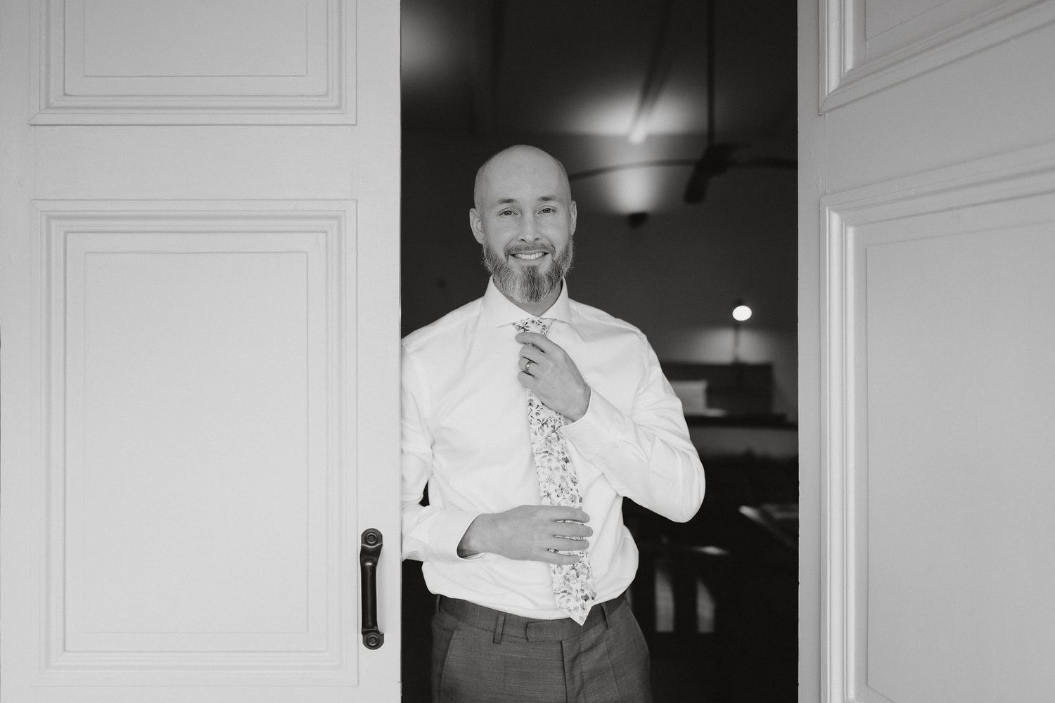 A person in a white dress shirt and tie adjusts their collar while standing in a doorway in this black and white photo.