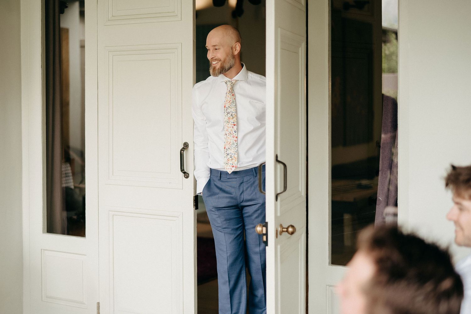 A person in a white dress shirt with floral tie and jeans stands in a doorway with white French doors.