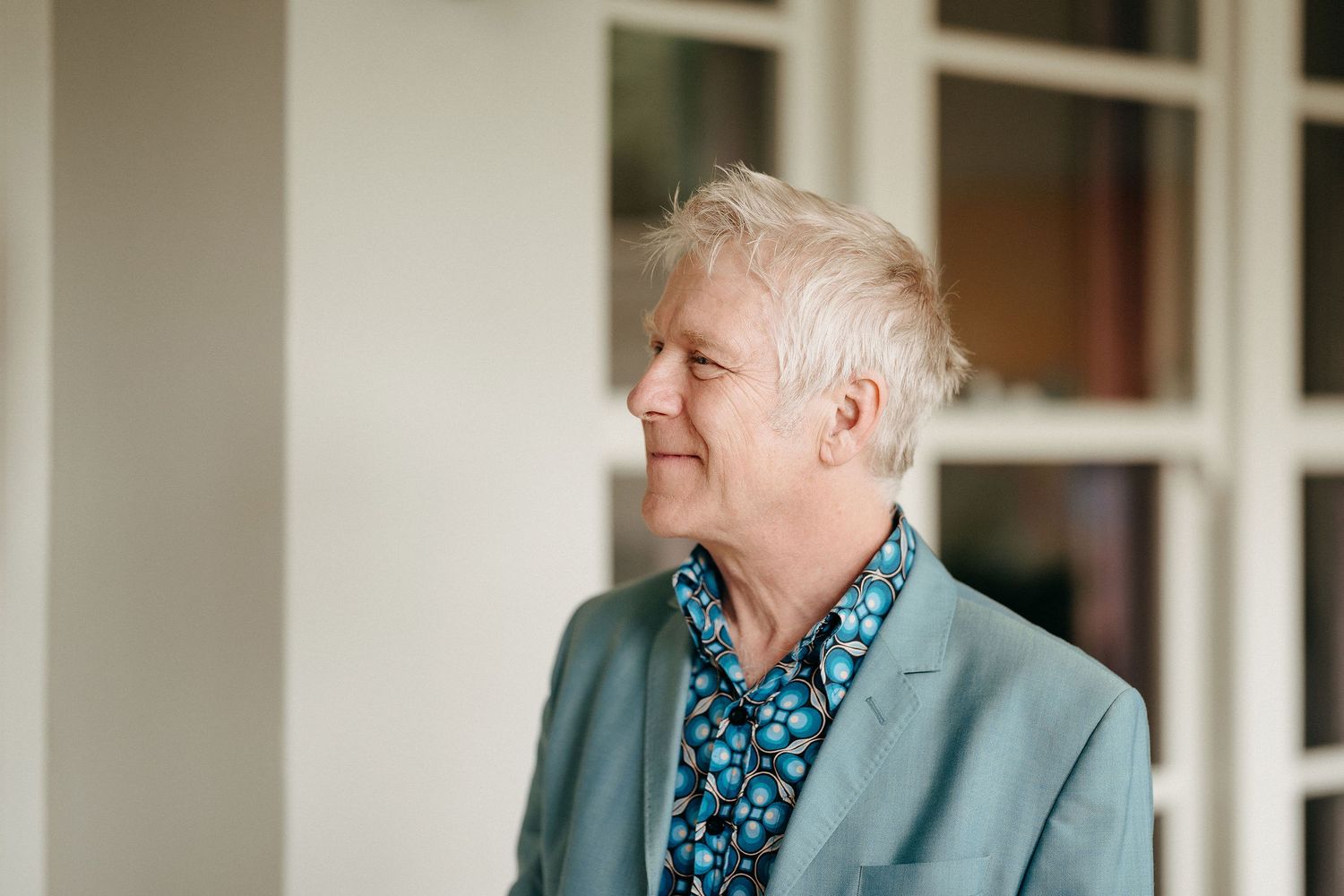 A person in a mint green blazer and floral shirt looks to the side while standing by a window.