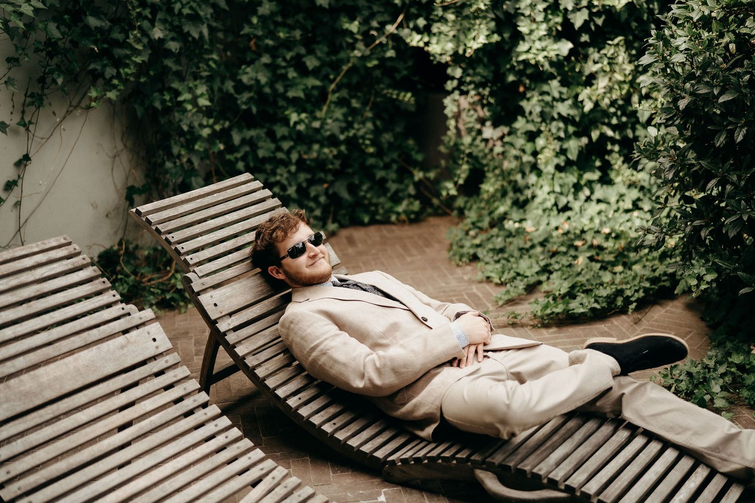 Person in light colored suit relaxes on curved wooden lounge chair surrounded by green foliage.