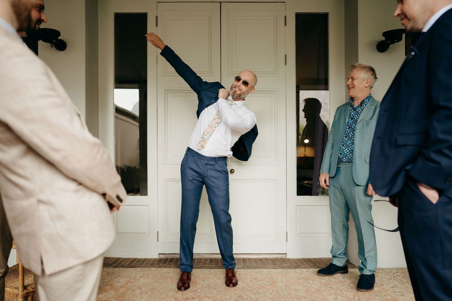 A person strikes an energetic dancing pose in front of a white doorway while others in suits look on.