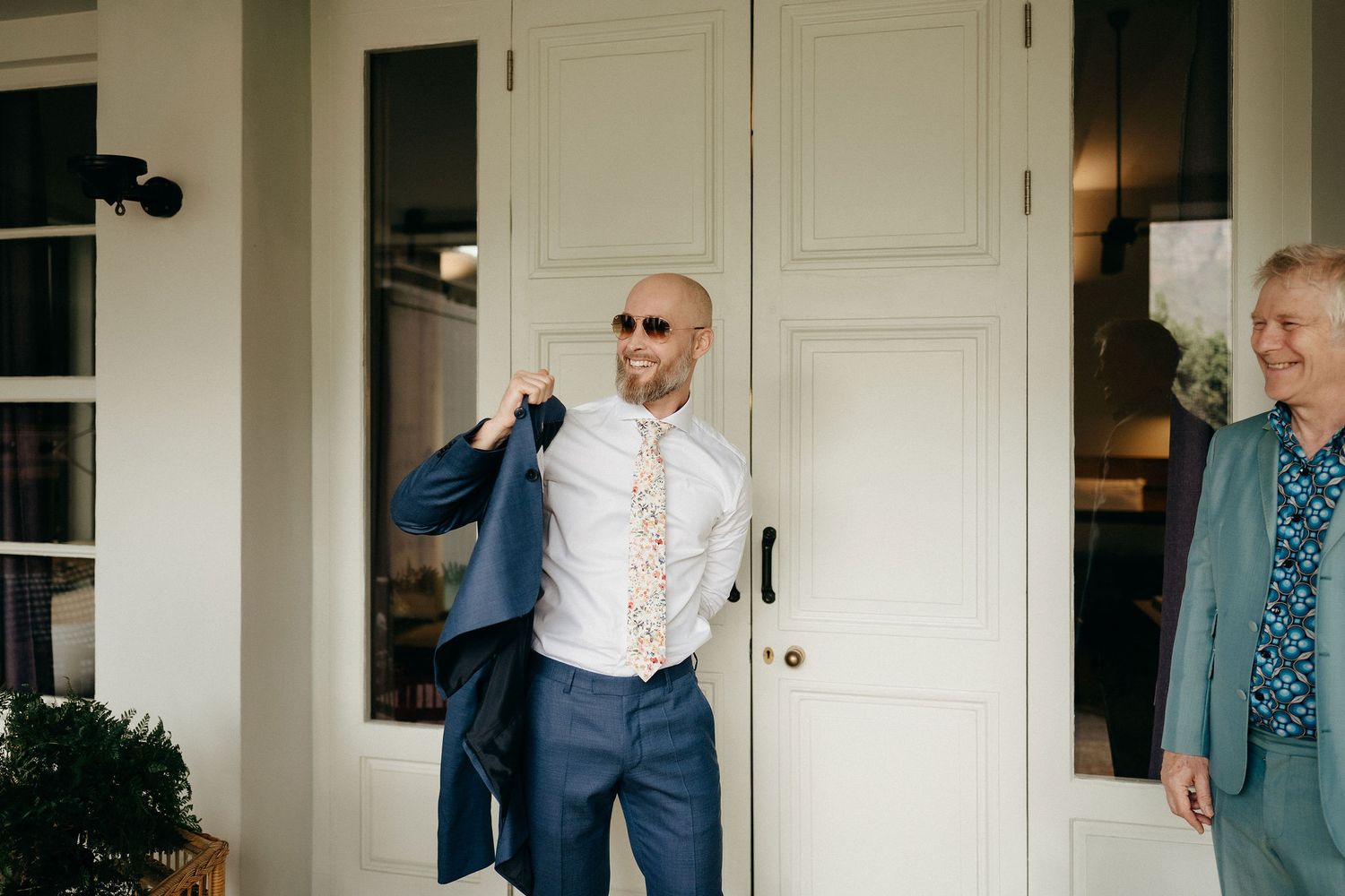 A person puts on a navy suit jacket while standing in front of white French doors at an elegant entrance.