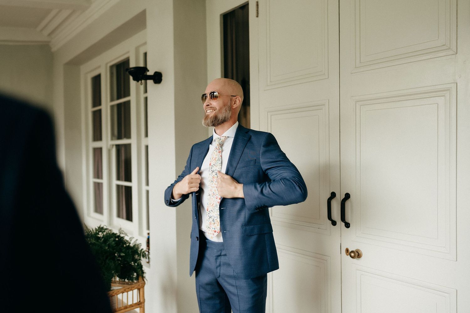 A person in a navy blue suit and tie adjusts their jacket while standing on a white porch.