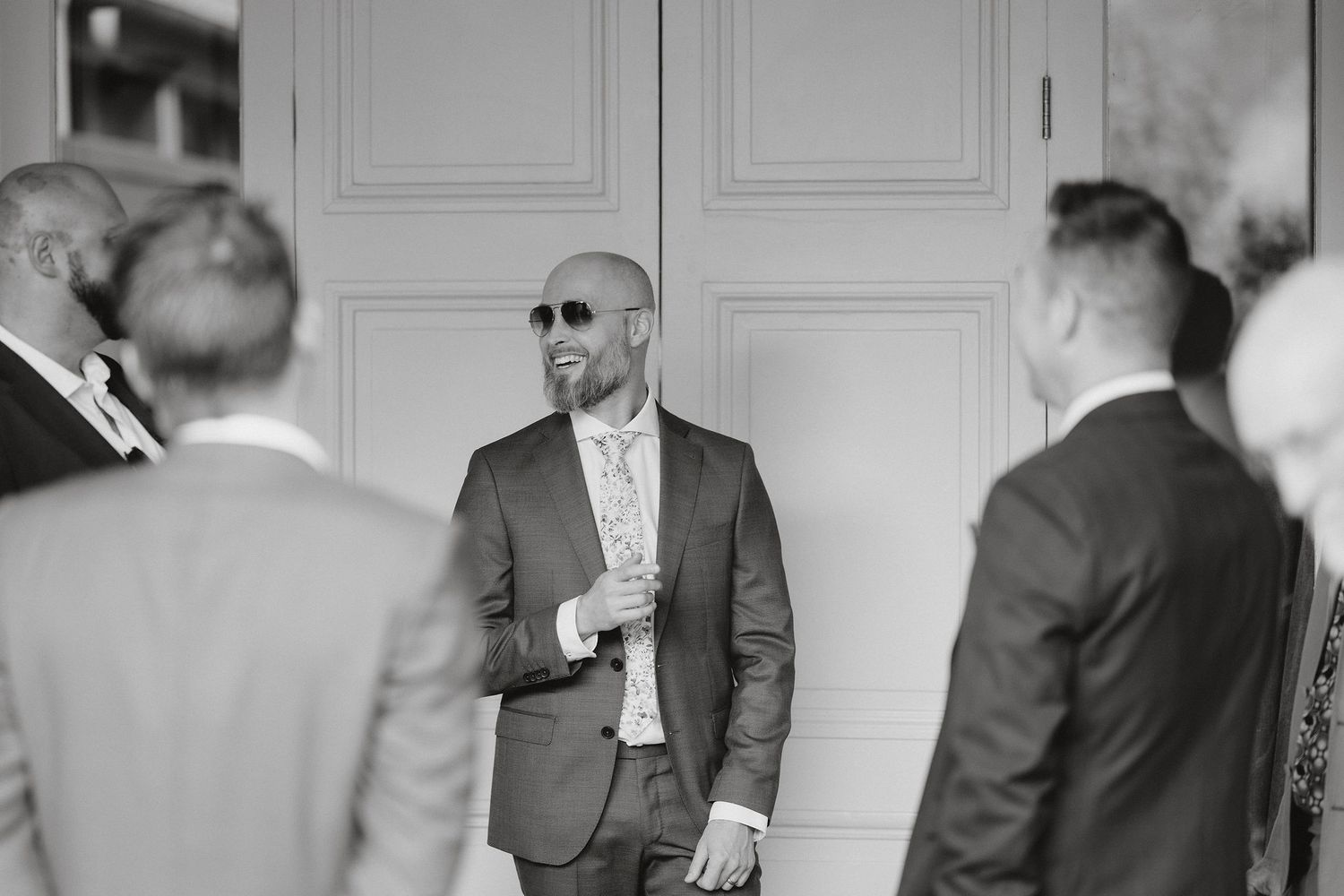 A person in sunglasses and a gray suit stands near white doors during what appears to be a formal gathering.