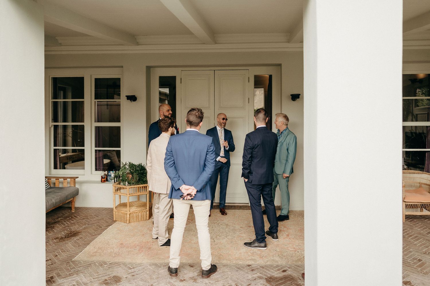 A group of people stand together on a front porch with white columns and French doors of an elegant home.
