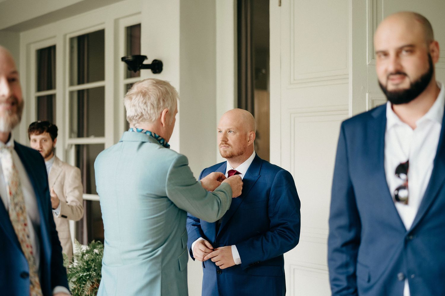 Men in formal attire converse on a white-columned porch during an elegant outdoor gathering.