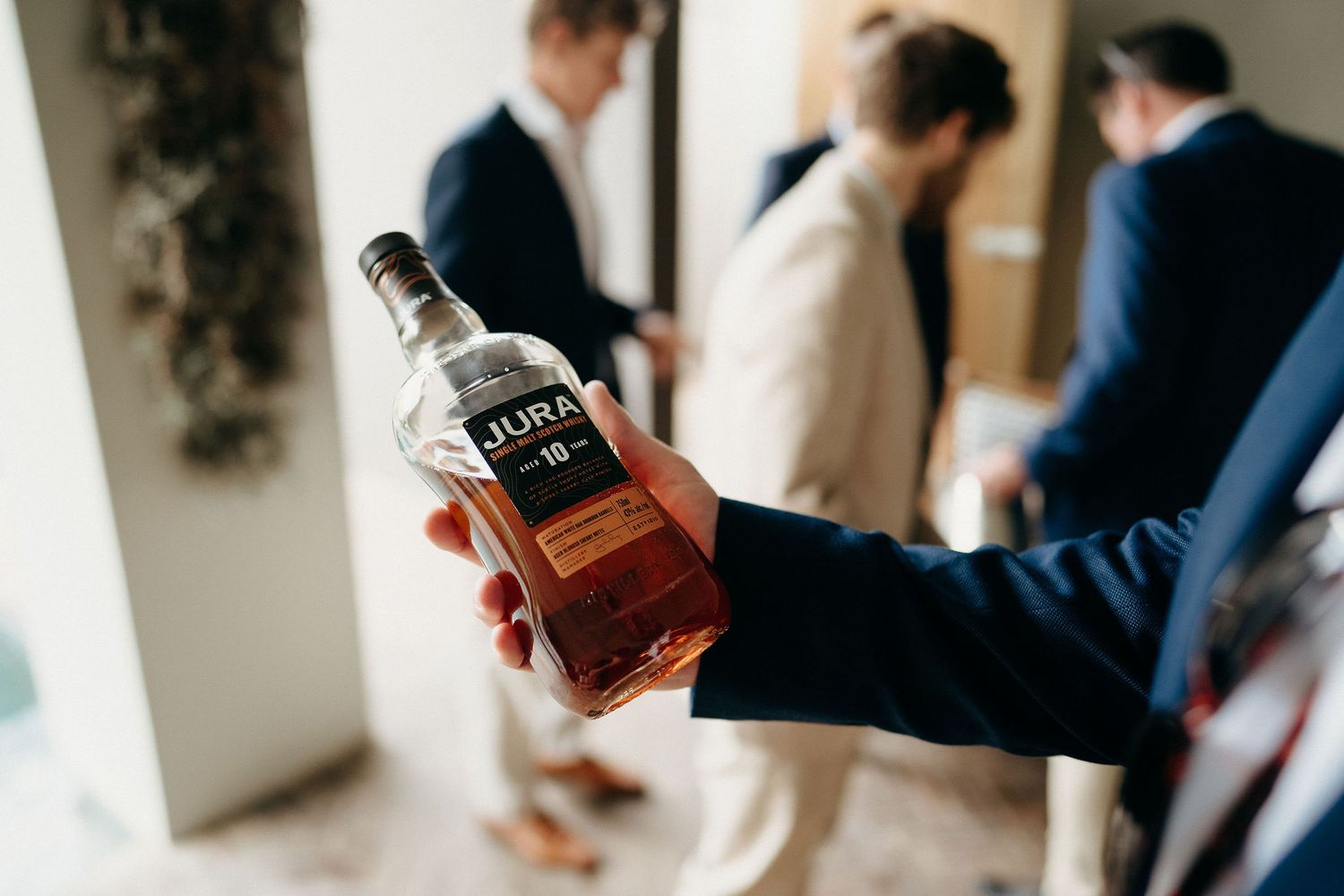 Hand holding a bottle of Jack Daniel's whiskey with people standing in the background of an indoor setting.