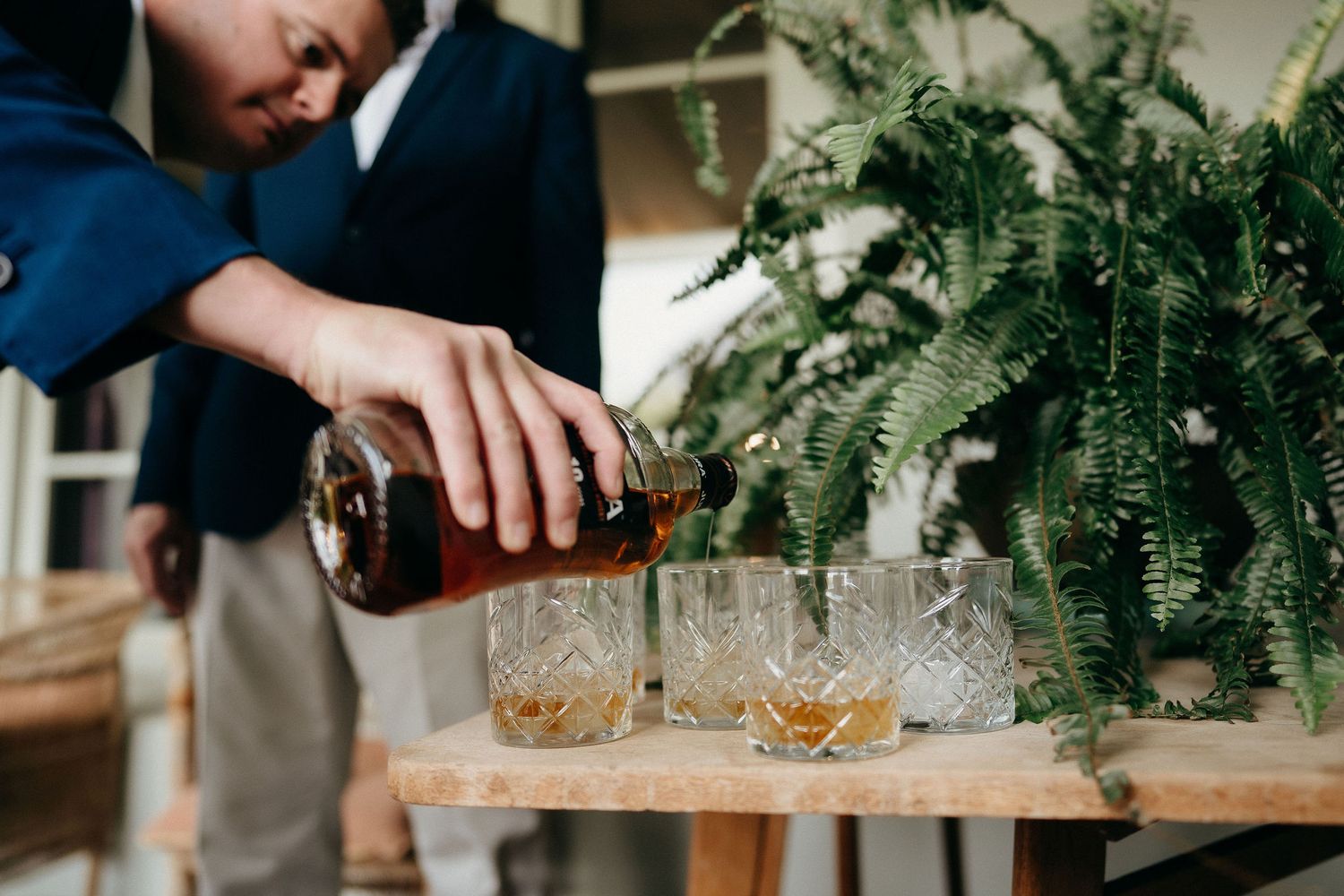 A person pours amber liquor into crystal glasses on a wooden table next to a lush green fern.