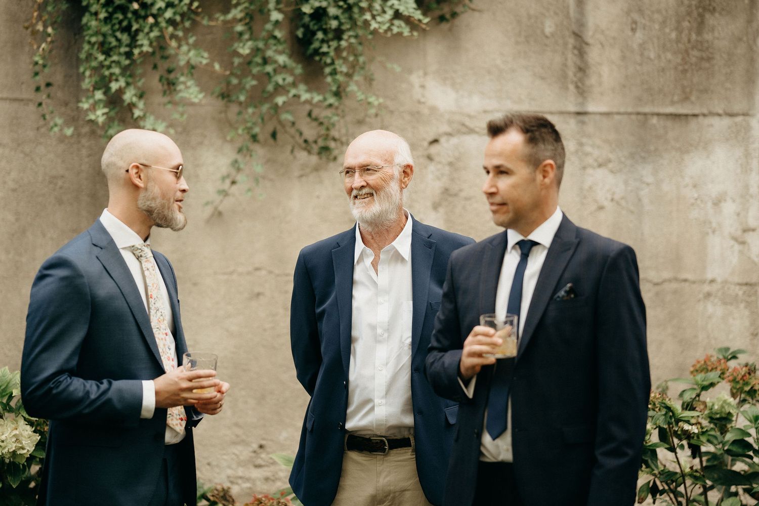 Three well-dressed men share drinks and conversation while standing outdoors against a stone wall with climbing vines.