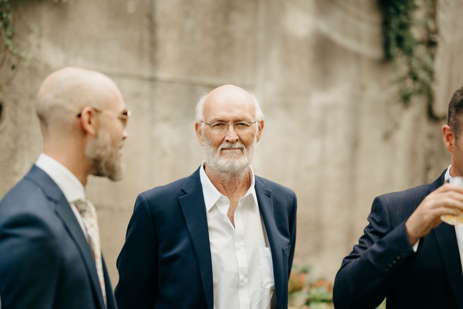 Three men in formal attire engaged in conversation outdoors against a concrete wall with hanging vines.