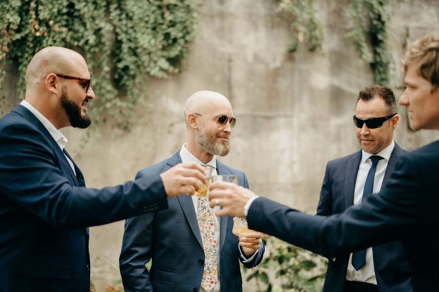 A group of men in suits raise their glasses in a celebratory toast against a wall covered with hanging vines.
