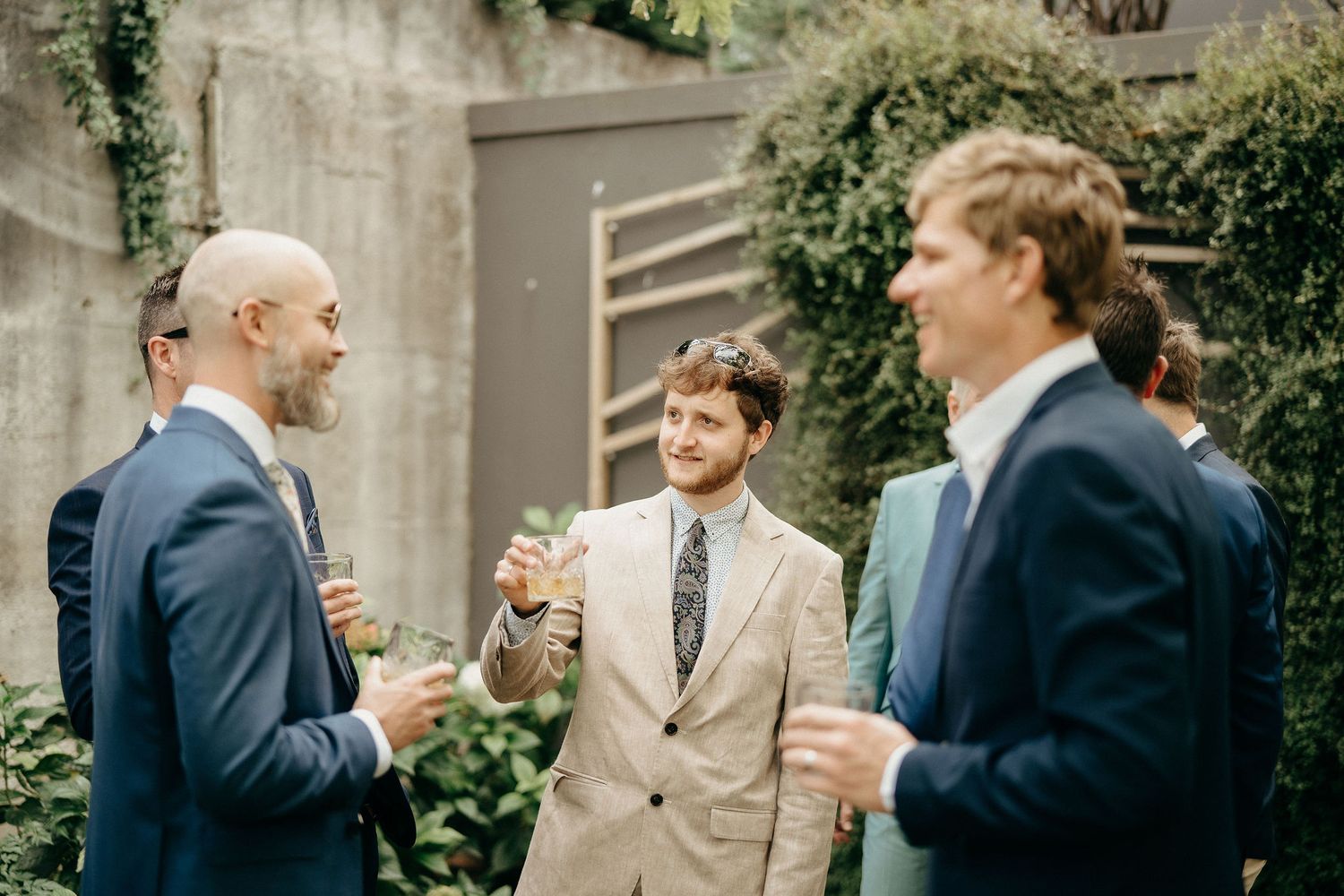Three men in suits share a lighthearted conversation while holding drinks at an outdoor gathering.