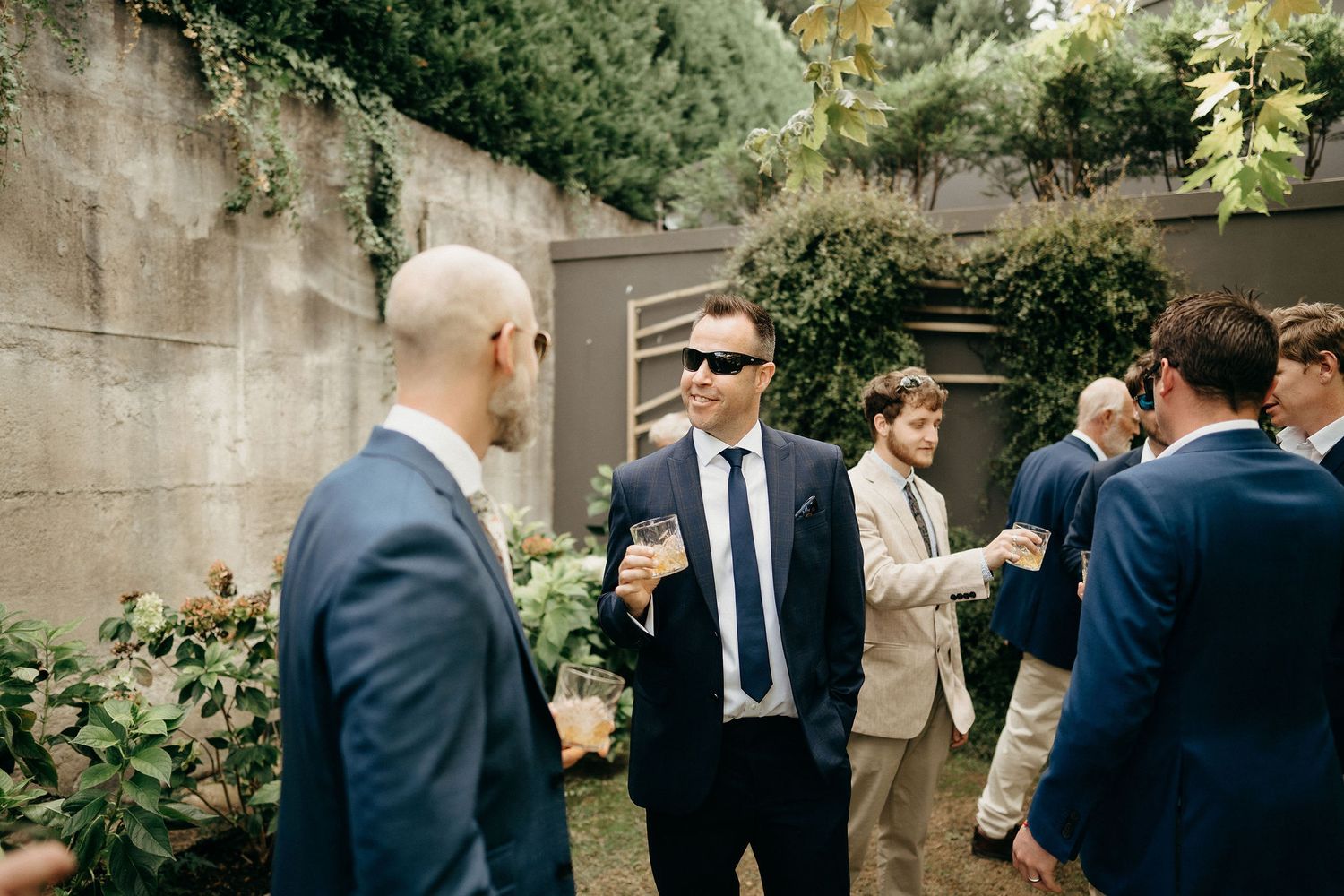 Guests in formal attire enjoying drinks and conversation in a garden setting with stone walls and greenery.