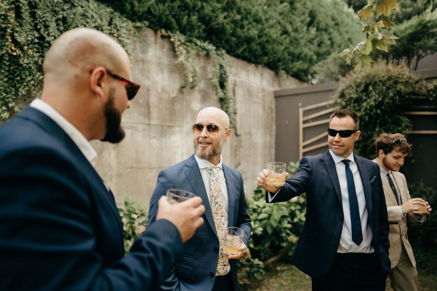 Group of well-dressed people in suits and sunglasses having a conversation outdoors by a stone wall.