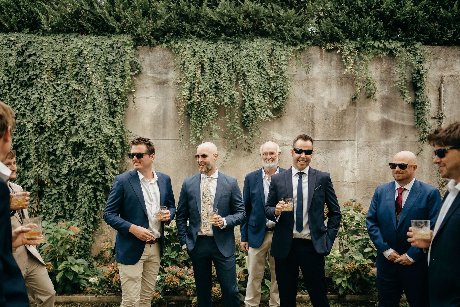 A group of well-dressed men in blue blazers and sunglasses stand casually together against an ivy-covered wall.