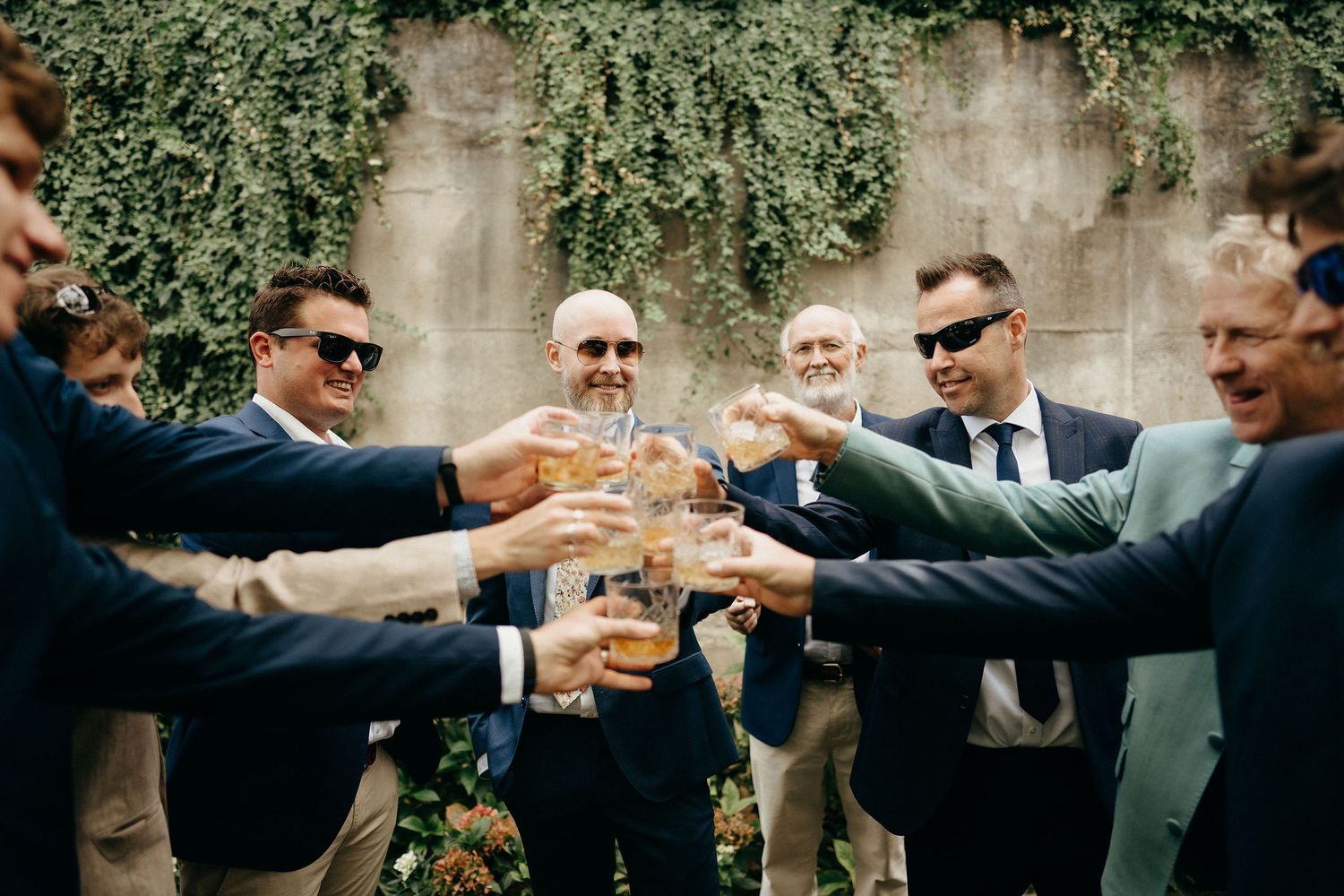 A group in formal suits gather in celebration to toast with drinks against an ivy-covered wall.