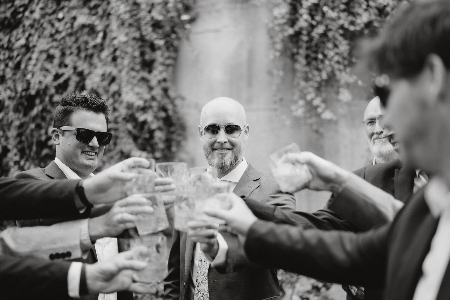 Group of people in formal attire making a celebratory toast with wine glasses at an outdoor event in black and white.