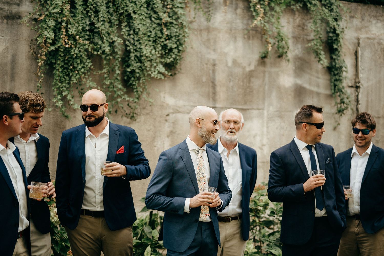 A group of stylishly dressed wedding guests stand against an ivy-covered wall holding drinks at an outdoor reception.