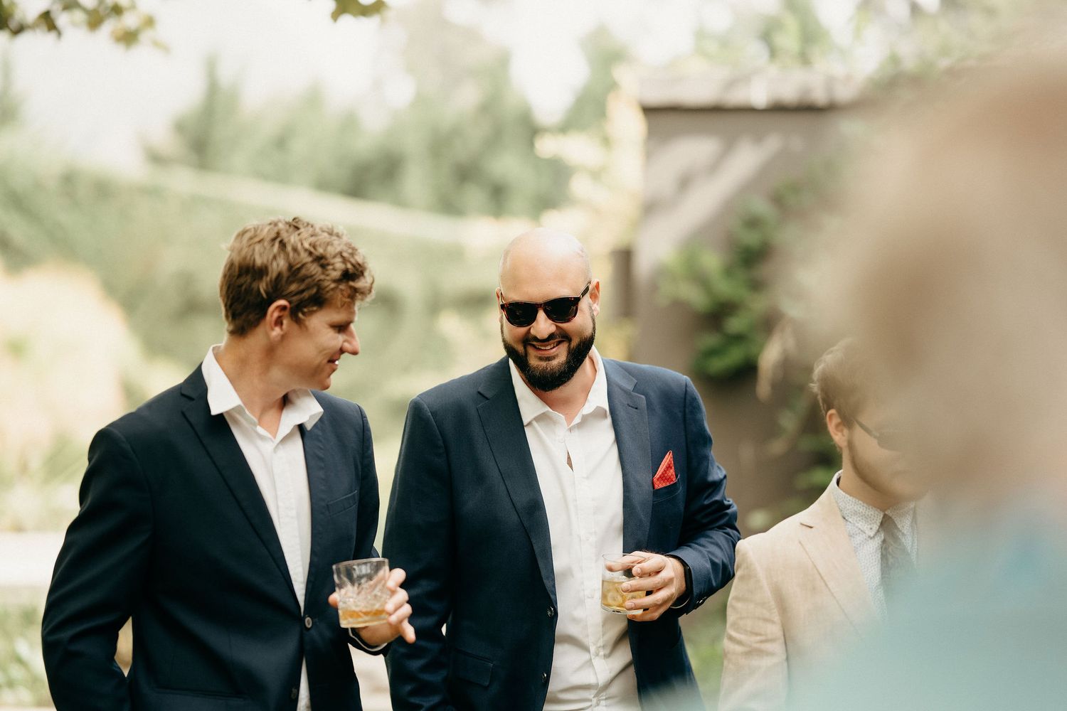 Two men in suits socialize at an outdoor event with soft bokeh lighting effects in the background.