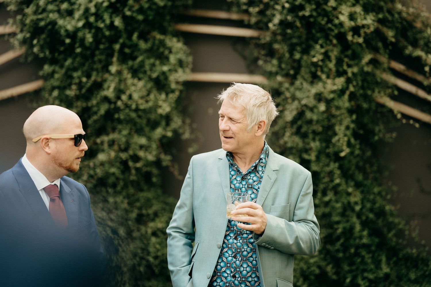 Two men stand outside, one wearing sunglasses and a suit, the other in a light blue blazer and patterned shirt.